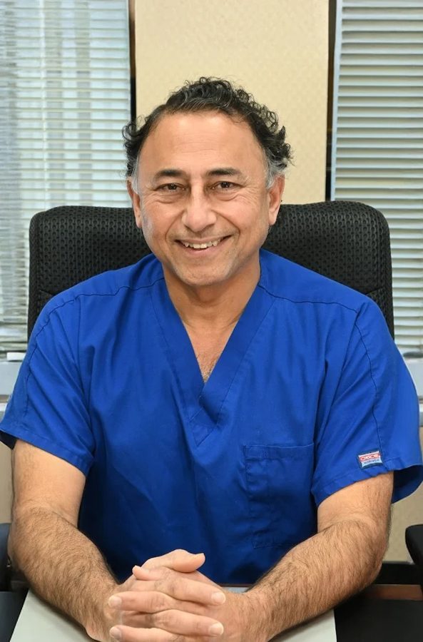 El Dr. Rajesh Khanna con uniforme quirúrgico azul, sonriendo en su escritorio en Khanna Vision Institute
